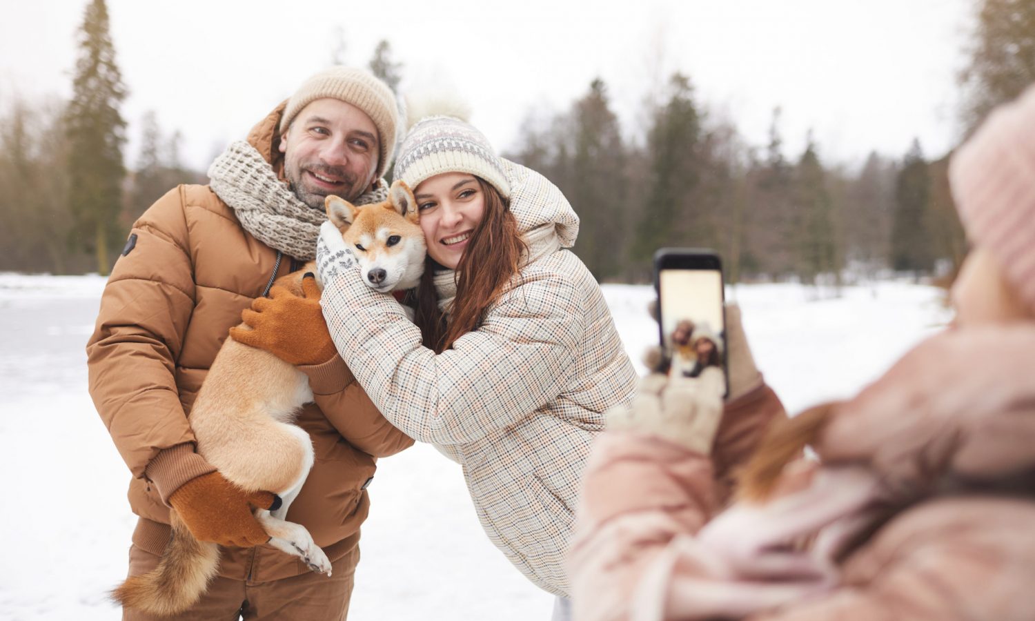 Teenage girl taking photo of parents while enjoying walk outdoors together in winter forest, focus on happy adult couple holding dog